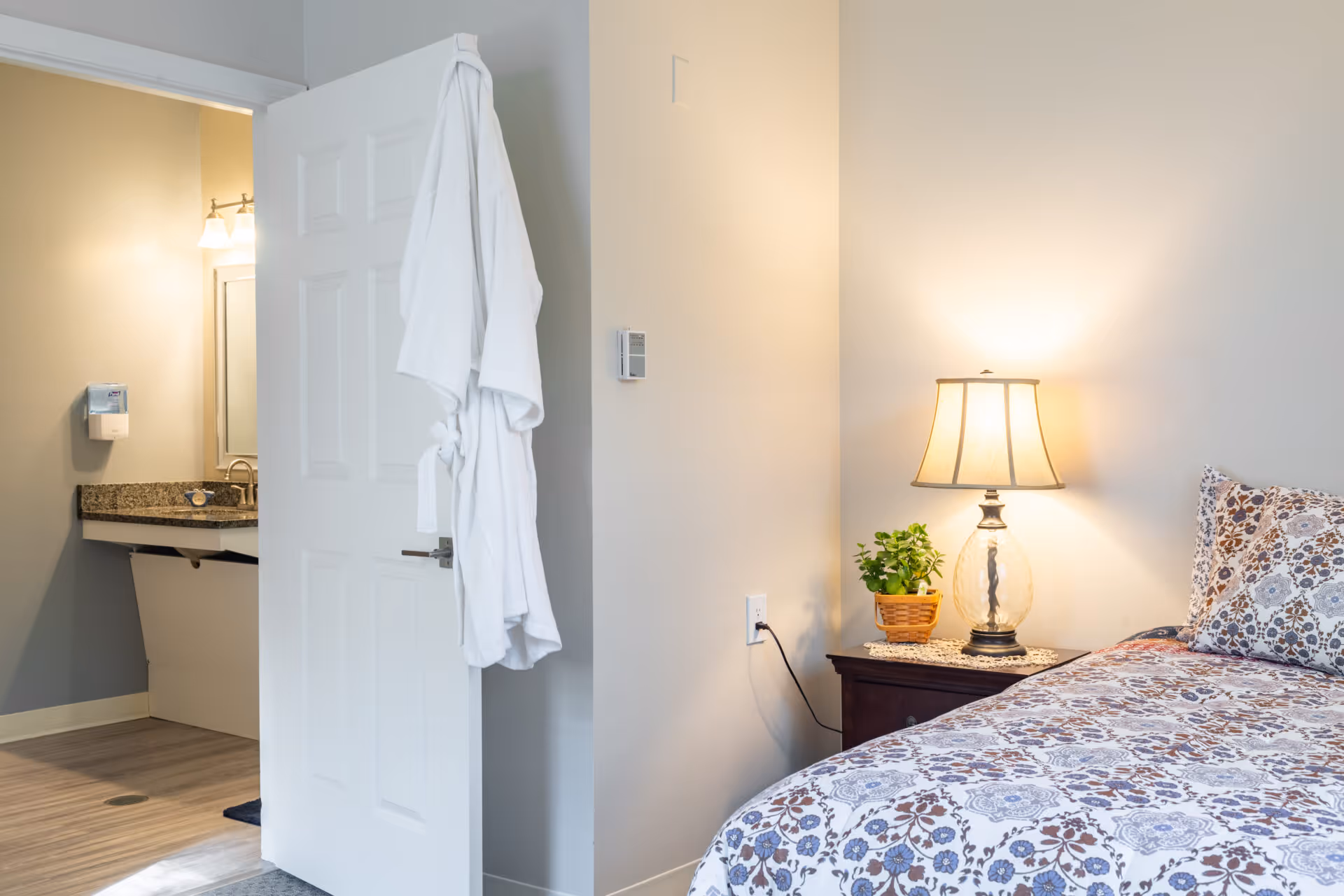 A cozy bedroom corner with a bed covered in a floral patterned bedspread and a matching pillow. Next to the bed is a wooden nightstand with a lit table lamp and a small potted plant. An open door reveals a bathroom sink area with a granite countertop, a mirror, and a white bathrobe hanging on the door.