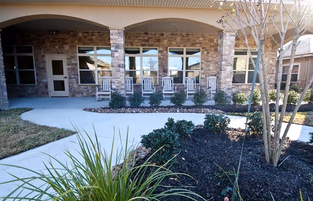 Outdoor patio area of a building with stone pillars and walls, featuring a row of white rocking chairs on a covered porch. In front of the porch is a curved concrete walkway surrounded by landscaped garden beds with bushes, plants, and a small tree.