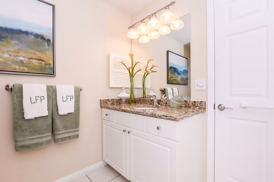 A clean bathroom vanity with a granite countertop, a sink, and a large mirror above it. Two green towels with white hand towels embroidered with 'LFP' hang on a towel rack on the wall. A vase with flowers and decorative glass items are placed on the countertop. A framed abstract painting is visible on the wall.
