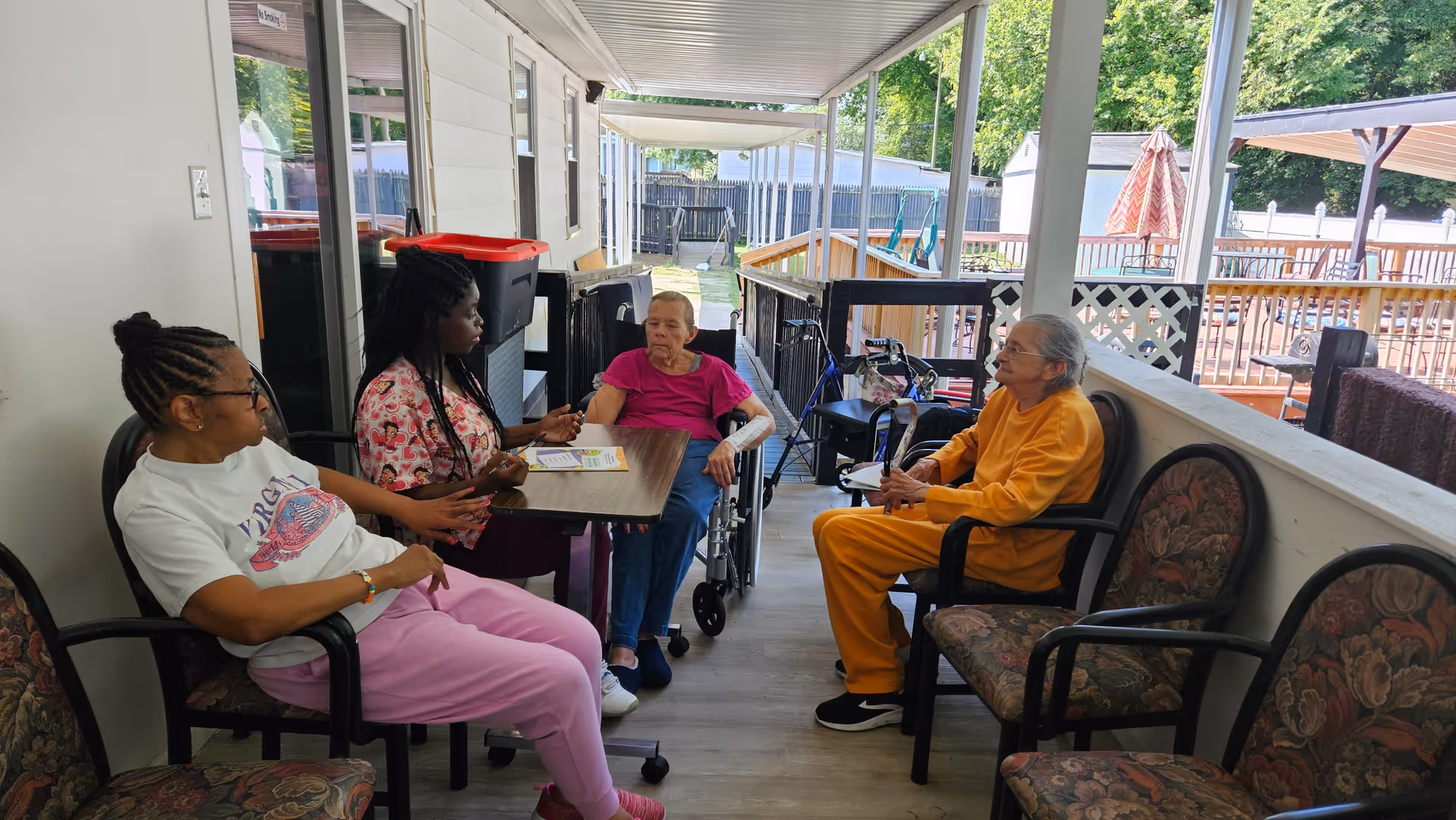 Four women sitting and conversing on a covered outdoor porch area with chairs and a table. One woman is in a wheelchair, another is wearing a bright orange outfit, and the setting overlooks a fenced yard with trees and outdoor furniture.
