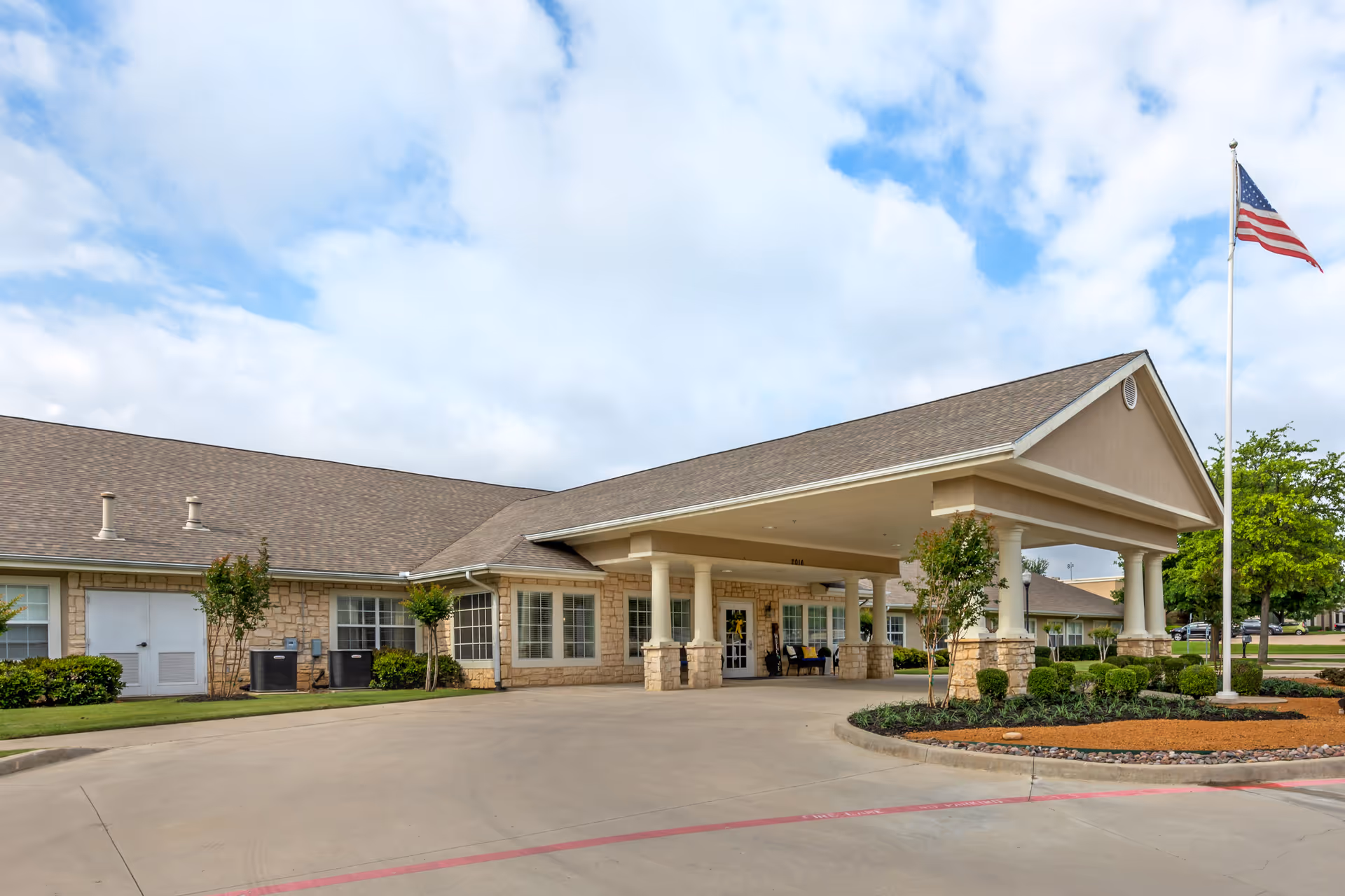 Exterior view of Brookdale Oak Hollow facility showing a single-story building with a covered entrance supported by columns, landscaped greenery, and an American flag on a flagpole under a partly cloudy sky.