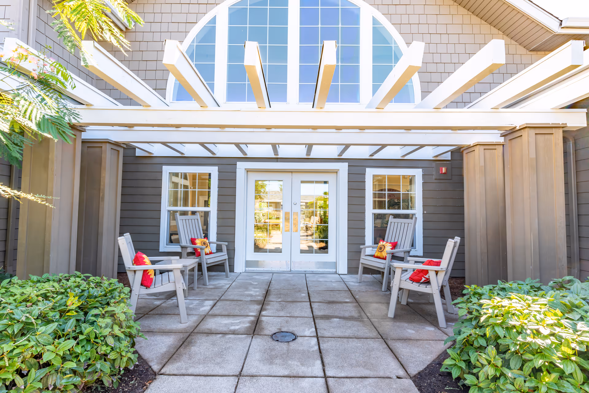 Front entrance with double glass doors under a white pergola, four outdoor chairs with colorful cushions, and surrounding shrubs.
