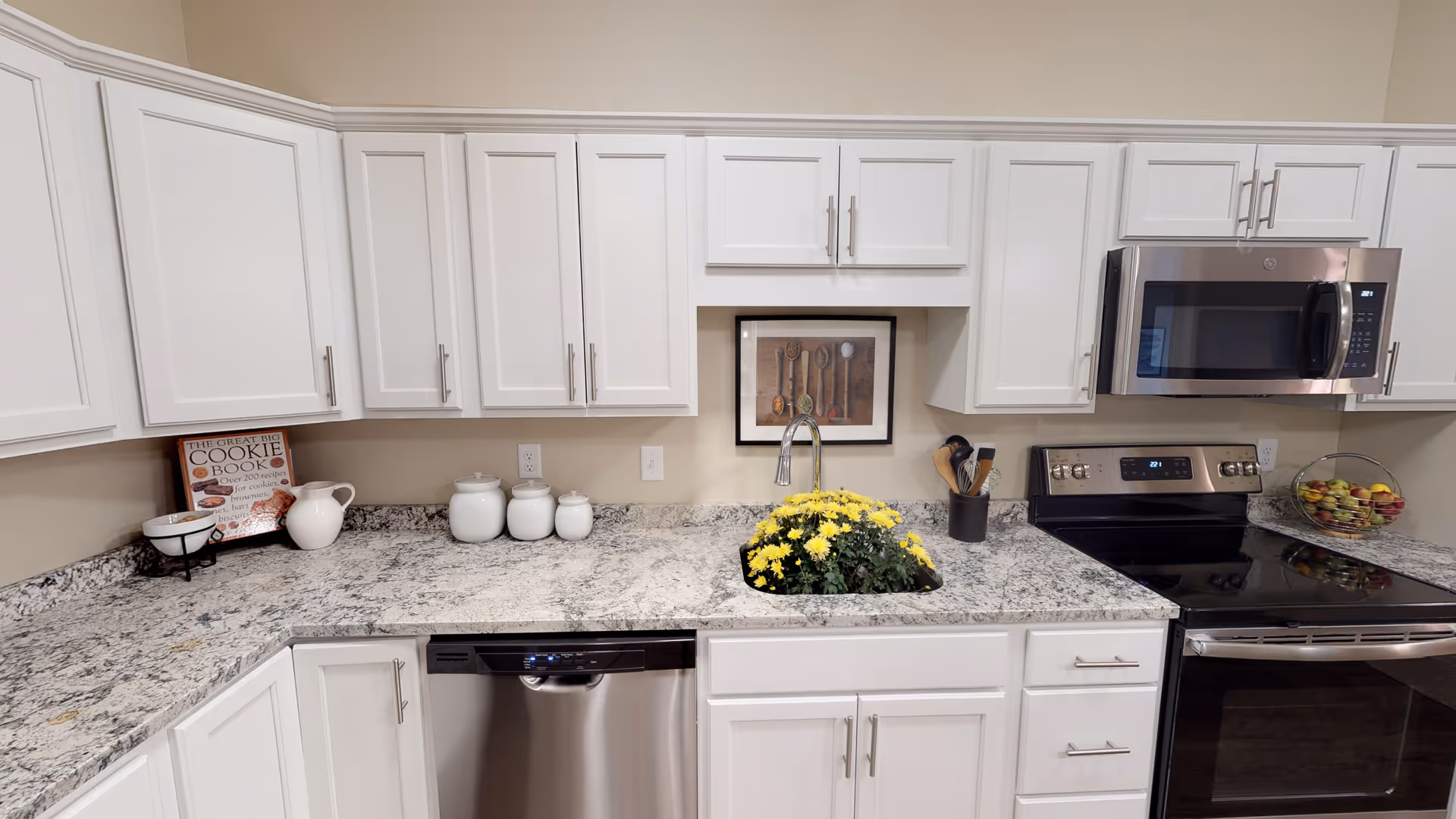 Modern kitchen with white cabinets, granite countertops, a stainless steel dishwasher, stove, and microwave. A sink with yellow flowers is centered under a framed picture of spoons. Various kitchen items including a cookie book, jars, and a fruit basket are placed on the countertops.