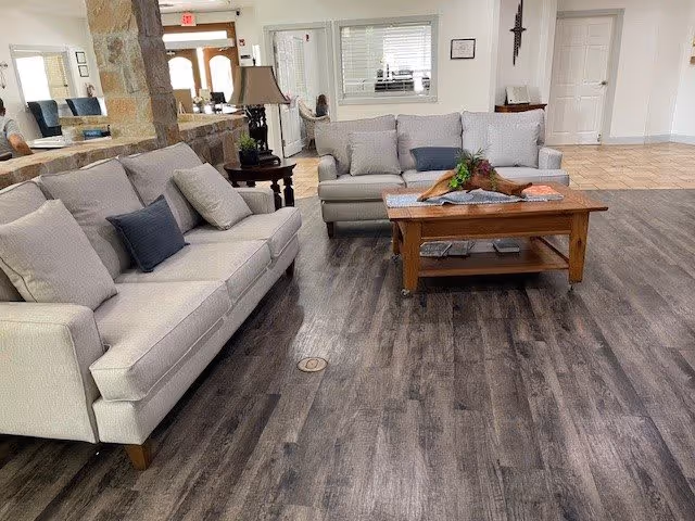 A cozy living room area with two light gray sofas facing each other, a wooden coffee table with decorative items on it, a side table with a lamp, and a large mirror on the wall reflecting part of the room. The floor is covered with dark wood-style flooring and the walls are painted white. There is a door and a window with blinds in the background.