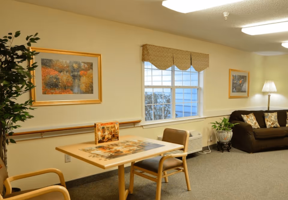 A cozy interior room with a small table and two chairs, a large window with a valance, a brown sofa with patterned cushions, a floor lamp, potted plants, and framed artwork on the walls.