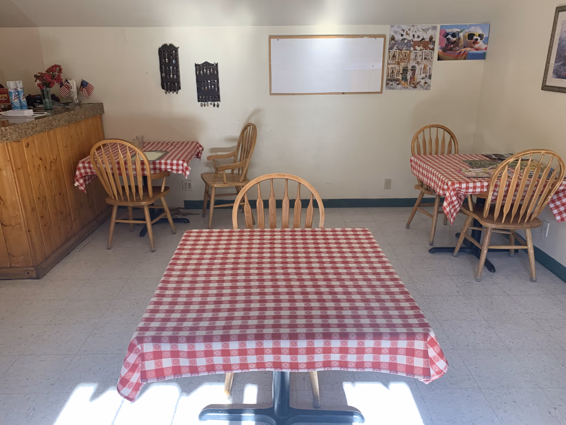 Small dining area with three wooden tables covered in red and white checkered tablecloths, each surrounded by wooden chairs. The room has a wooden counter on the left side with some items and small American flags on it. The walls are decorated with a whiteboard, two small hanging displays, and pictures including one of dogs wearing sunglasses.