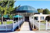 Covered circular entrance canopy and walkway leading to the front of a single-story nursing facility with white picket fences and lawn.