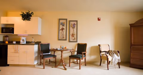Interior view of a senior living facility room featuring a small kitchenette with white cabinets, a microwave, and a coffee maker on the left. In the center, there is a round wooden table with two green cushioned chairs. On the right, there is a wooden armoire and an additional chair with a light-colored throw draped over it. Two framed floral artworks hang on the wall above the table.
