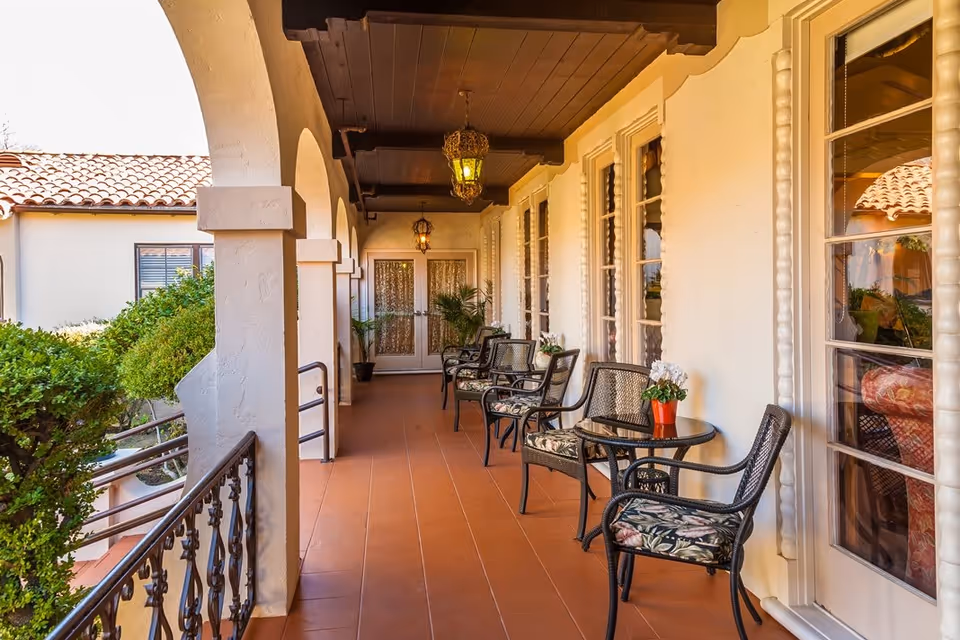 A covered outdoor patio area with terracotta tile flooring and white stucco walls. The patio features black metal chairs with floral cushions and small round tables with potted plants. Arched openings overlook greenery and a tiled roof building. Decorative hanging lanterns are suspended from the dark wooden ceiling.