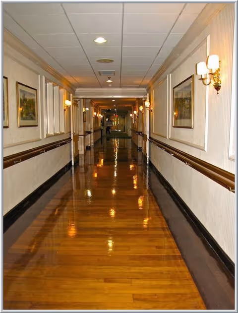A long, polished wooden hallway in a facility with handrails on both sides, wall-mounted lamps, framed paintings, and a drop ceiling with recessed lighting.