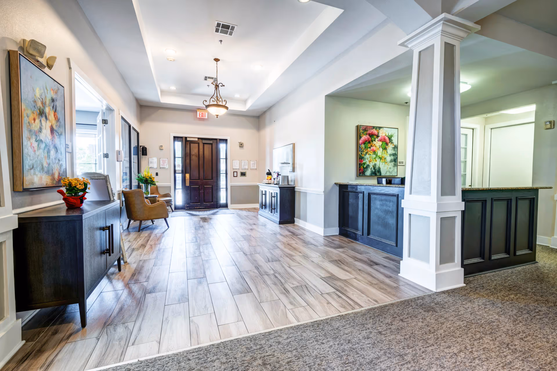 Bright and welcoming assisted living facility lobby with wood-look tile flooring, a dark wooden front door with sidelights, two brown armchairs near the door, a dark wooden sideboard with a flower vase and artwork on the wall, a chandelier hanging from the ceiling, and a reception desk with a granite countertop and floral painting behind it.