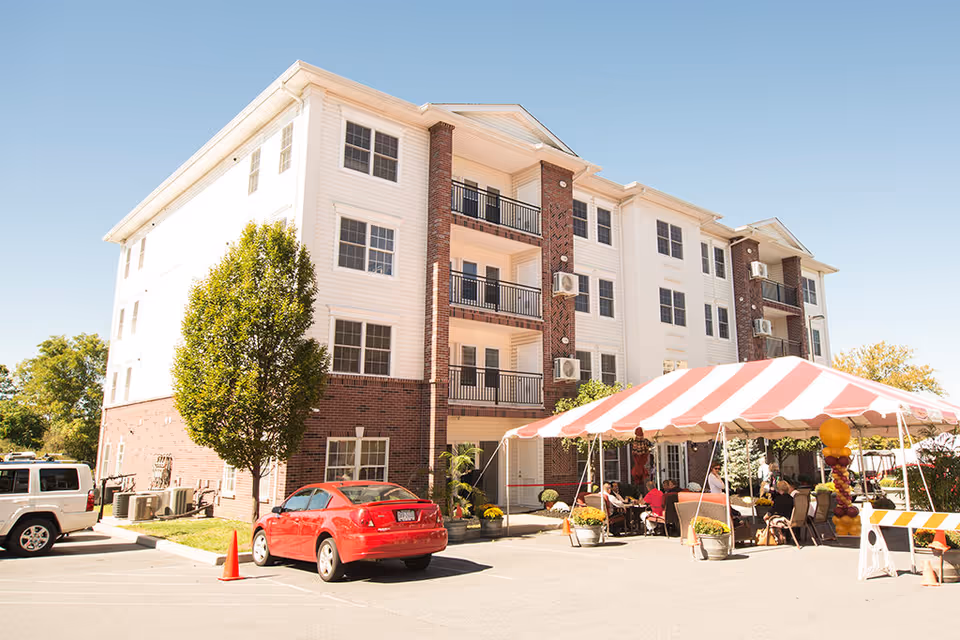 Exterior view of a multi-story senior living facility building with brick and white siding. There is a red and white striped tent set up outside with people sitting underneath it. Several cars are parked in front of the building, and there are trees and plants around the area under a clear blue sky.