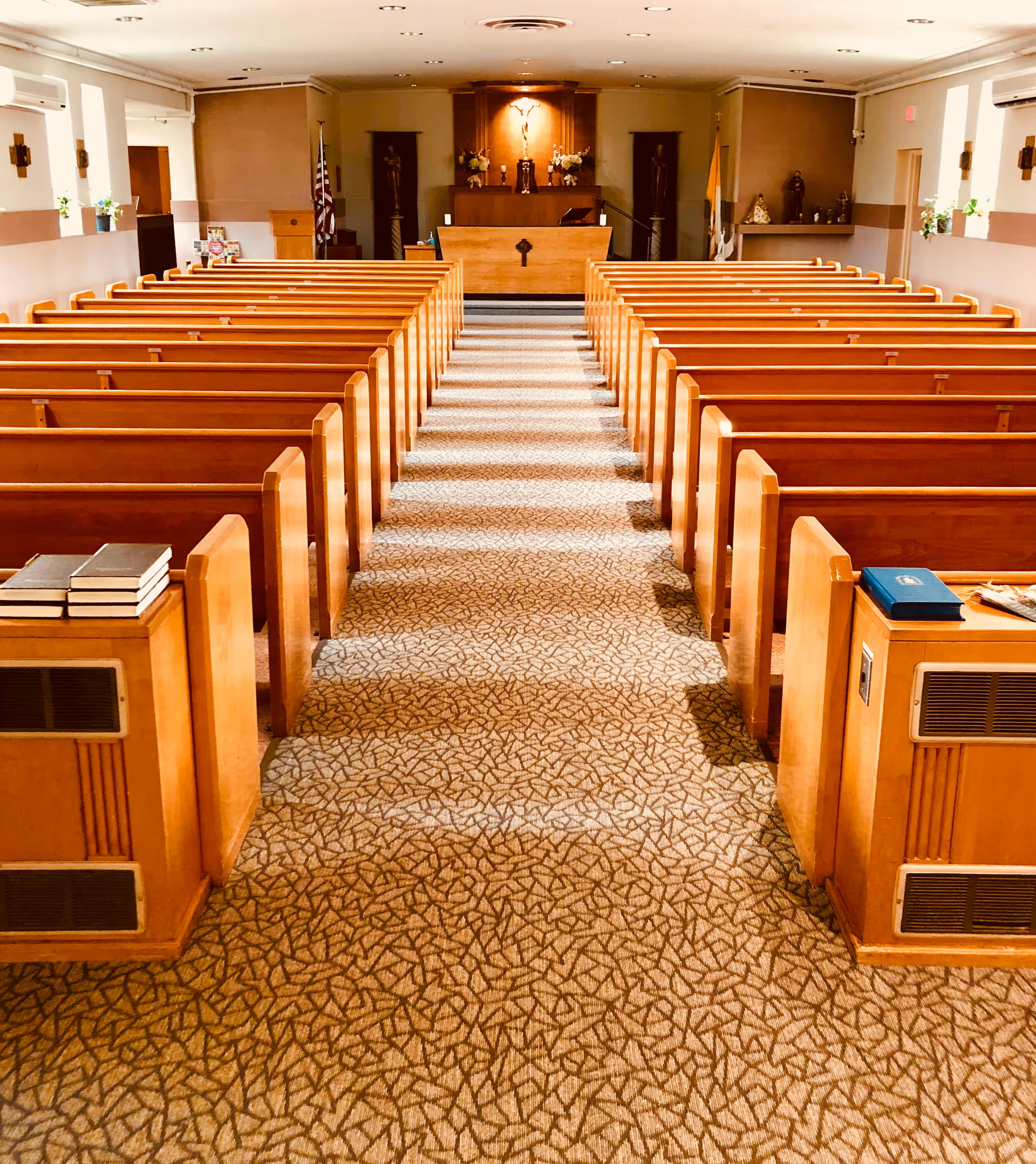 Interior view of a chapel or worship room with wooden pews arranged in rows on both sides of a central carpeted aisle leading to an altar at the front. The altar area features religious statues, flowers, and flags on either side. The room is softly lit with ceiling lights.