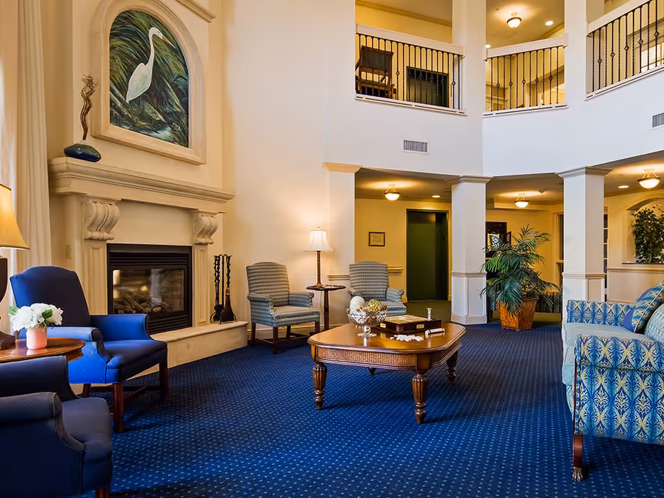 A furnished senior living lounge with blue carpet, a fireplace, armchairs, a central coffee table and an open two-story balcony.