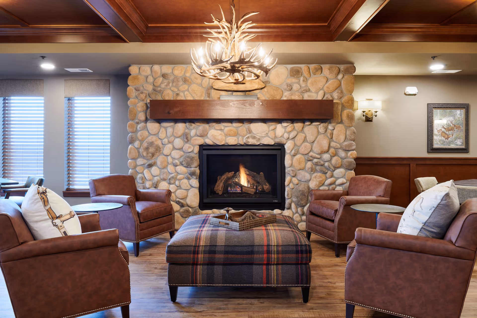 Cozy lounge with a stone fireplace, four leather armchairs around a plaid ottoman, and an antler chandelier.