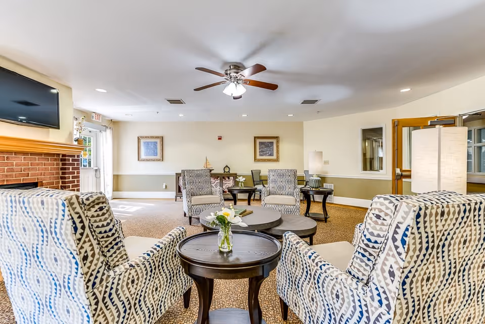 A bright and spacious living room with patterned armchairs arranged around a round wooden coffee table with a small vase of flowers. The room features a ceiling fan with lights, a brick fireplace with a mounted flat-screen TV above it, framed artwork on the walls, and a door leading outside. There are additional tables and chairs in the background, along with lamps and windows letting in natural light.