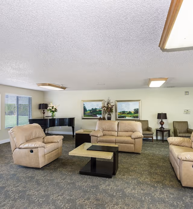A spacious living room with beige leather sofas and armchairs arranged around two nested coffee tables. The room has a patterned carpet, two framed landscape paintings on the wall, a black grand piano with a vase of flowers on top, and two table lamps on side tables. Large windows with blinds allow natural light into the room.