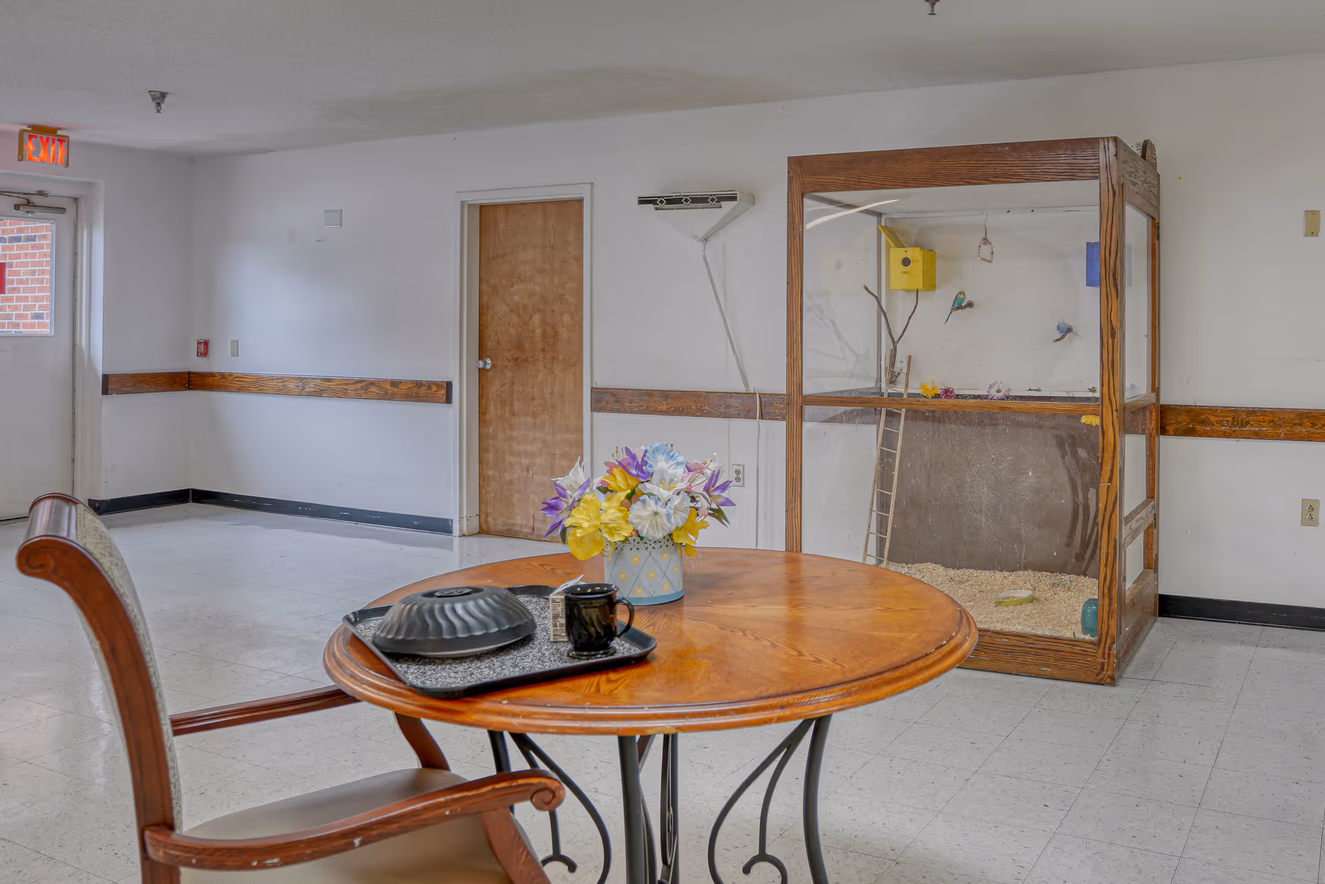 A small seating area in a hallway or common space with a round wooden table and a single wooden chair. On the table is a tray with a covered dish, a black mug, and a small container, along with a vase of colorful artificial flowers. In the background, there is a large wooden and glass birdcage with two small birds inside. The walls are white with a wooden chair rail, and there is a closed wooden door and an exit door with a red exit sign above it.