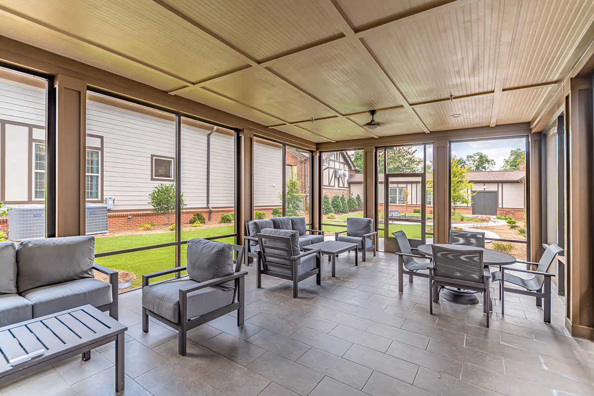 A screened-in patio area with cushioned outdoor seating including sofas and armchairs, a coffee table, and a round dining table with chairs. The patio overlooks a well-maintained garden with grass, shrubs, and neighboring buildings.