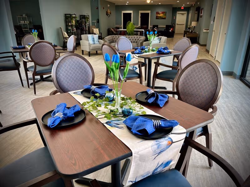 A dining room in a senior living facility with multiple tables set for four people each. The tables have dark wood surfaces, black plates with blue cloth napkins, and a floral table runner with a vase of blue tulips and green leaves as centerpiece. The room has light-colored flooring, upholstered chairs, and a seating area with sofas and armchairs in the background.