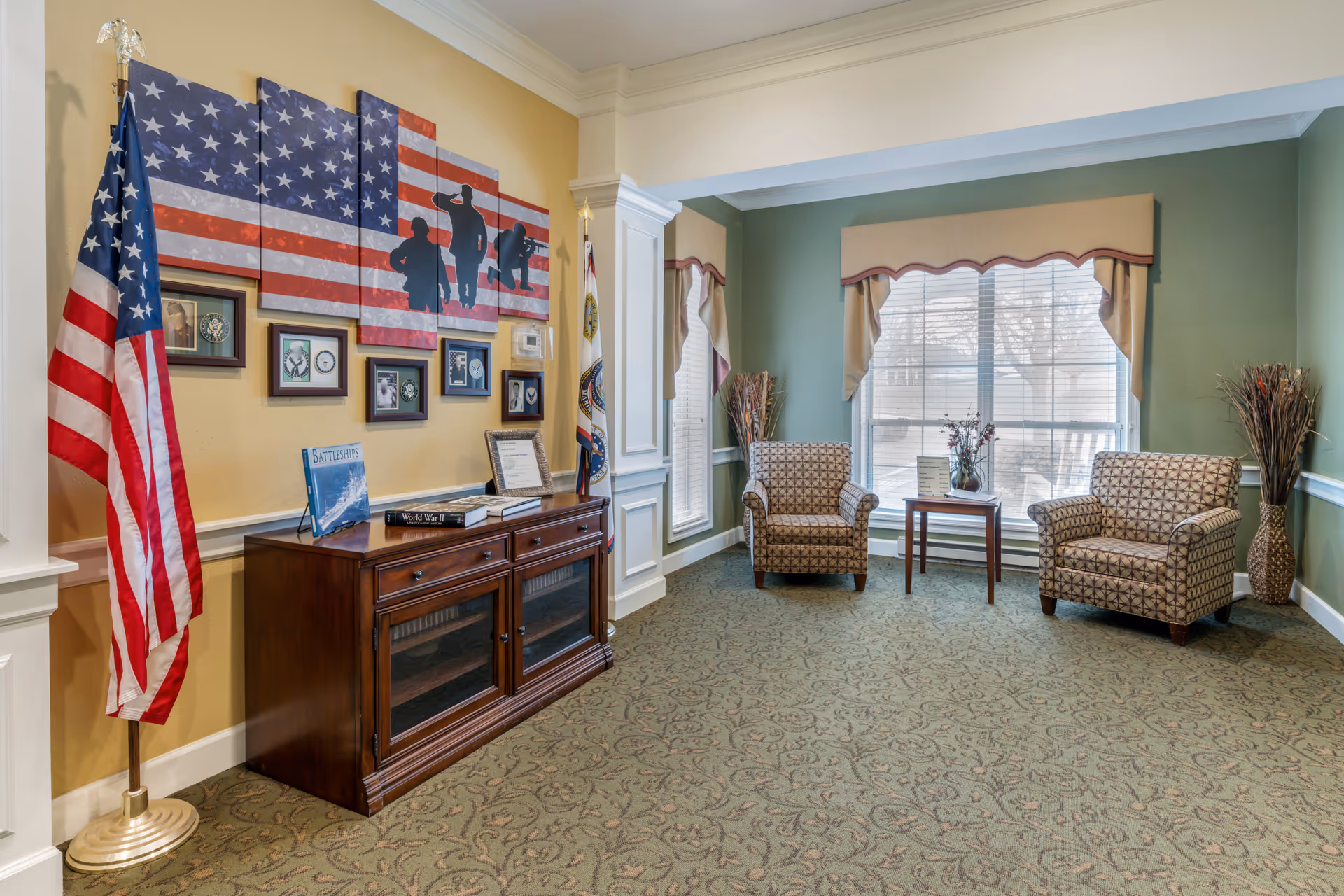 A cozy sitting area in a senior living facility with two patterned armchairs and a small wooden table between them, placed in front of a large window with beige valances. To the left, there is a wooden cabinet with books and framed pictures above it, including a large American flag artwork with silhouettes of soldiers. An American flag and another flag stand on either side of the cabinet. The walls are painted in soft yellow and green tones, and the floor is covered with a patterned carpet.