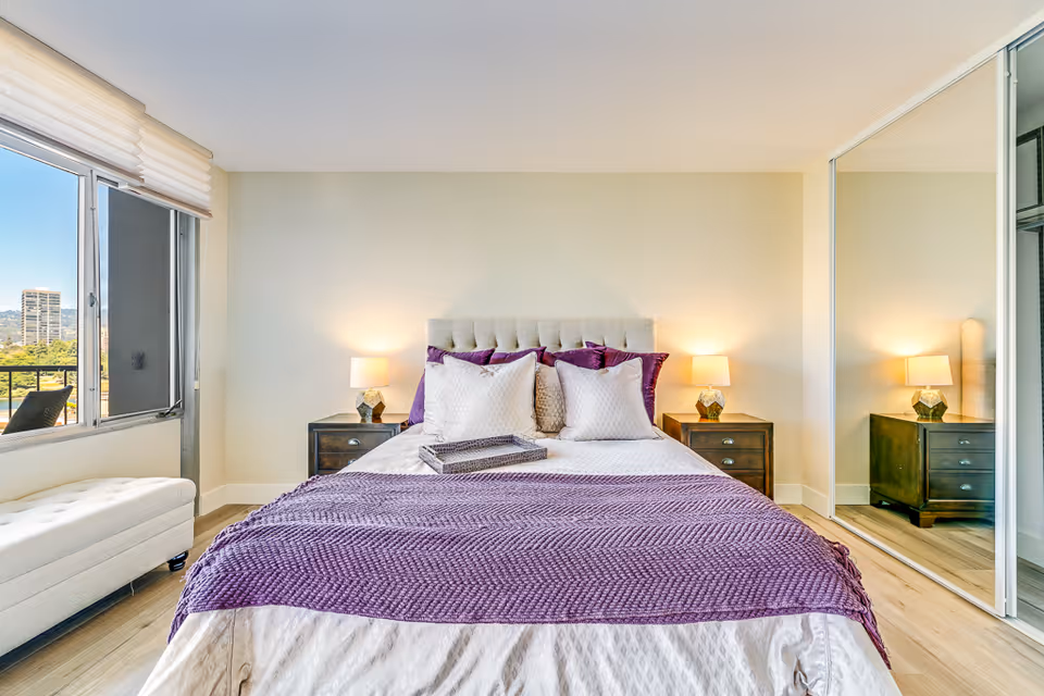 A bright bedroom with a large bed centered against a light-colored wall. The bed is made with white and purple bedding, including a purple throw blanket and several pillows. There are two dark wooden nightstands on either side of the bed, each with a lamp. To the right, there is a large mirrored closet door reflecting part of the room. On the left, a window with a view of buildings and greenery outside lets in natural light. A white cushioned bench is placed under the window.
