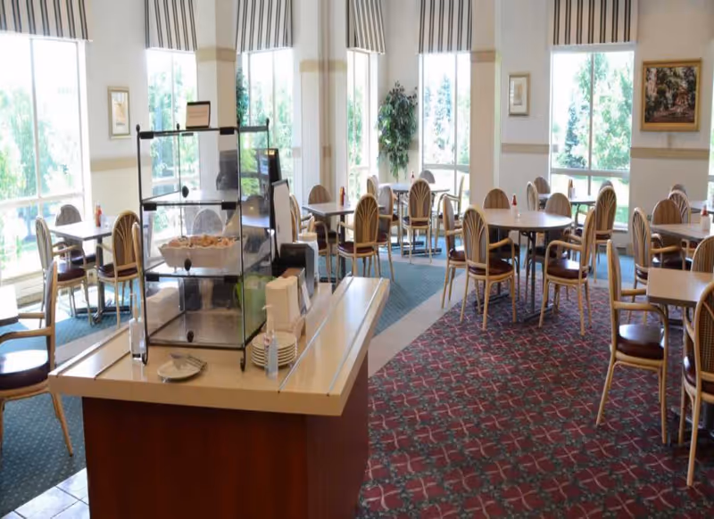 Bright dining room with round tables and chairs, a central buffet island holding pastries and plates, and large windows letting in daylight.