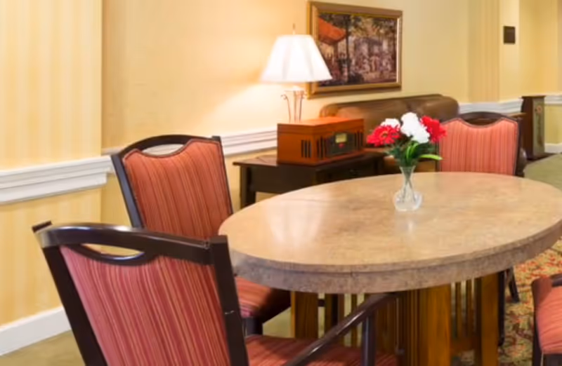 Oval dining table with a small vase of flowers surrounded by upholstered chairs in a warm, furnished common area.