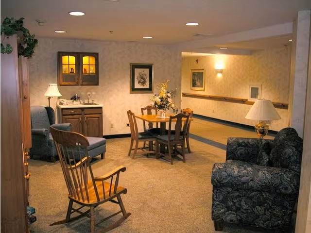A cozy senior living community common area featuring a wooden rocking chair, a small round table with four chairs, a floral centerpiece, a patterned sofa, an armchair, a wooden cabinet with a lamp, and framed artwork on the walls. The space is warmly lit with recessed ceiling lights and wall sconces.