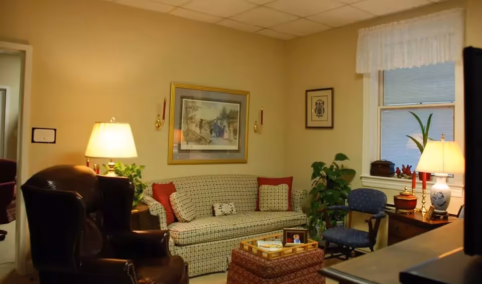 A cozy living room with a patterned sofa adorned with red and beige cushions, a dark leather armchair, a blue upholstered chair, and a wooden side table with a lamp and decorative items. There are framed pictures on the beige walls, a window with white curtains, and a potted plant near the window.