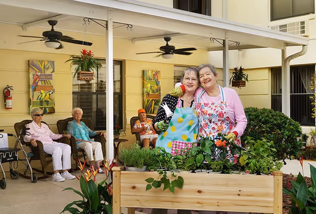 Two elderly women wearing aprons and gardening gloves smile and pose together behind a raised garden bed filled with plants. In the background, three other elderly individuals sit on chairs under a covered patio attached to a yellow building, with ceiling fans and hanging plants visible.