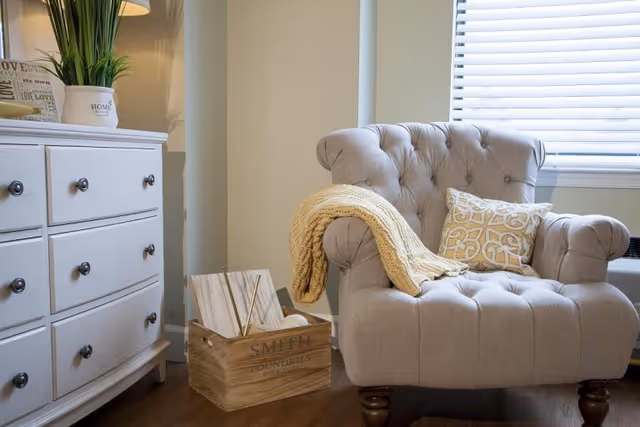 A cozy living room corner featuring a tufted beige armchair with a yellow knitted throw draped over one arm and a decorative patterned pillow. Next to the chair is a wooden crate labeled 'SMITH FOUNDRIES' holding some books. To the left is a white dresser with black knobs, topped with a potted plant and a lamp. A window with white blinds is partially visible behind the chair.