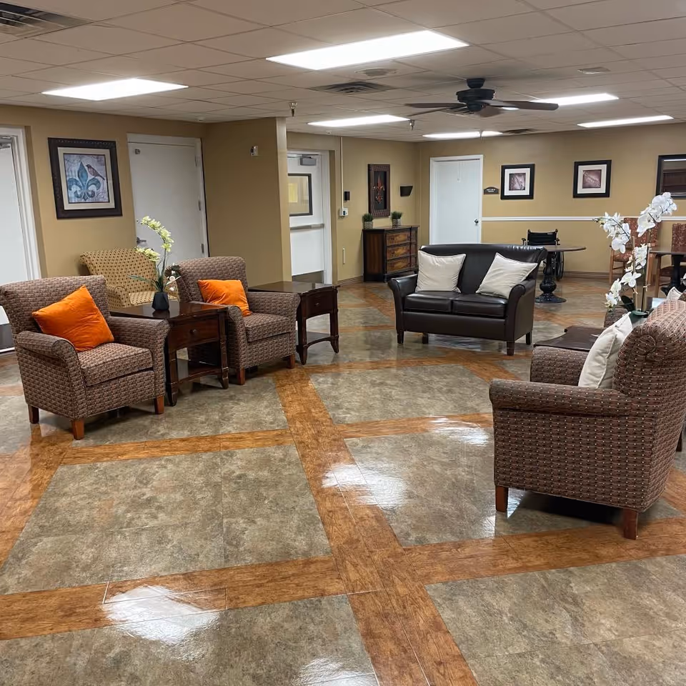 A spacious and well-lit common area in a healthcare facility featuring several armchairs with orange and white cushions, a dark brown loveseat, side tables, and decorative plants. The room has a tiled floor with a wood pattern, beige walls adorned with framed artwork, and a ceiling fan.