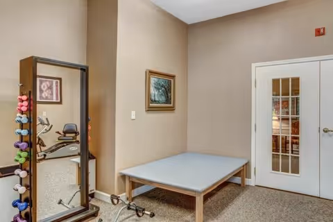 A small exercise room with beige walls and carpeted floor. There is a large mirror with colorful dumbbells hanging on its side, a padded exercise platform, and a stationary exercise bike visible through an open doorway. A framed picture hangs on the wall above the exercise platform, and a set of white double doors with glass panes is on the right side of the room.