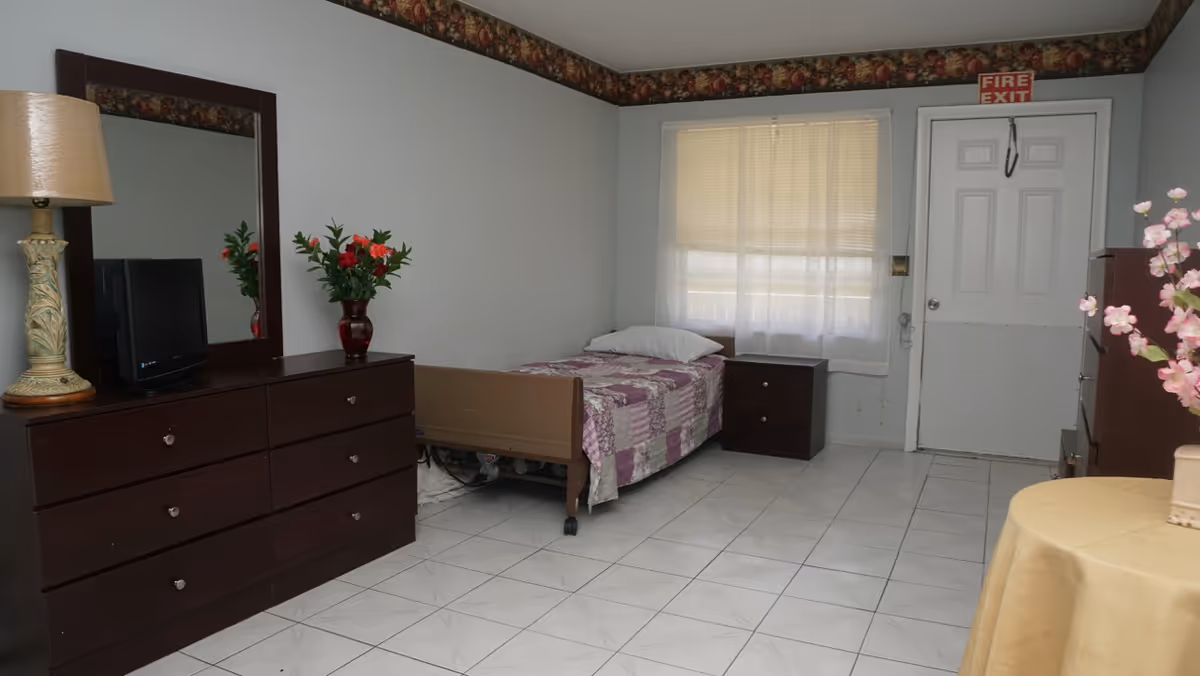 A simple bedroom with a single bed covered with a purple and white patchwork quilt, a pillow, and a dark wooden nightstand next to it. There is a large dresser with a mirror above it, a table lamp, a vase with flowers, and a small TV on top. The room has white tiled floors, light gray walls with floral wallpaper border near the ceiling, a window with blinds, and a white door with a fire exit sign above it. A round table with a yellow tablecloth and a vase with pink flowers is partially visible on the right.