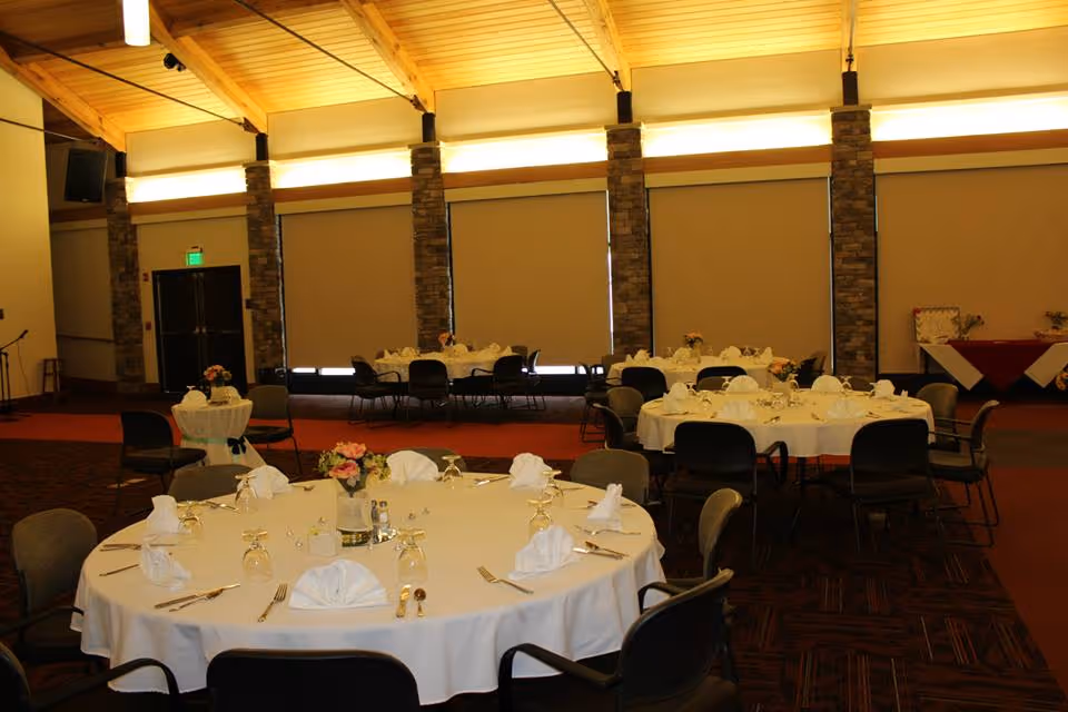 A spacious dining room with round tables covered in white tablecloths, each set with folded napkins, glassware, silverware, and floral centerpieces. The room features tall stone pillars, large windows with shades drawn, and a high wooden ceiling with warm lighting.