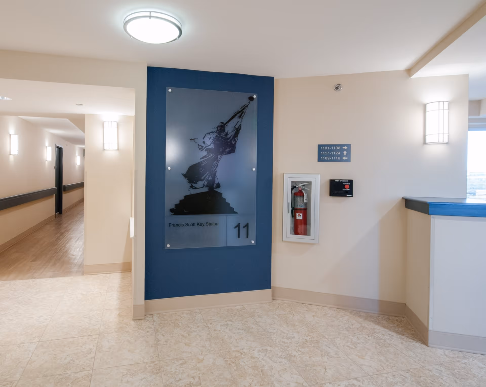 Interior hallway area of Linden Park Apartments in Bolton Hill with beige tiled floor, cream walls, and a blue accent wall featuring a glass panel with an image of the Francis Scott Key Statue and the number 11. There is a fire extinguisher mounted on the wall, directional signs for room numbers, and modern wall-mounted light fixtures illuminating the space.