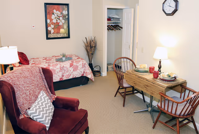 A cozy senior living room featuring a red armchair with a chevron-patterned pillow and a red throw blanket, a single bed with a floral bedspread, a small wooden dining table set for two with plates, cups, and napkins, a wall clock, a framed floral painting, a floor lamp, and an open closet with hangers and folded linens.