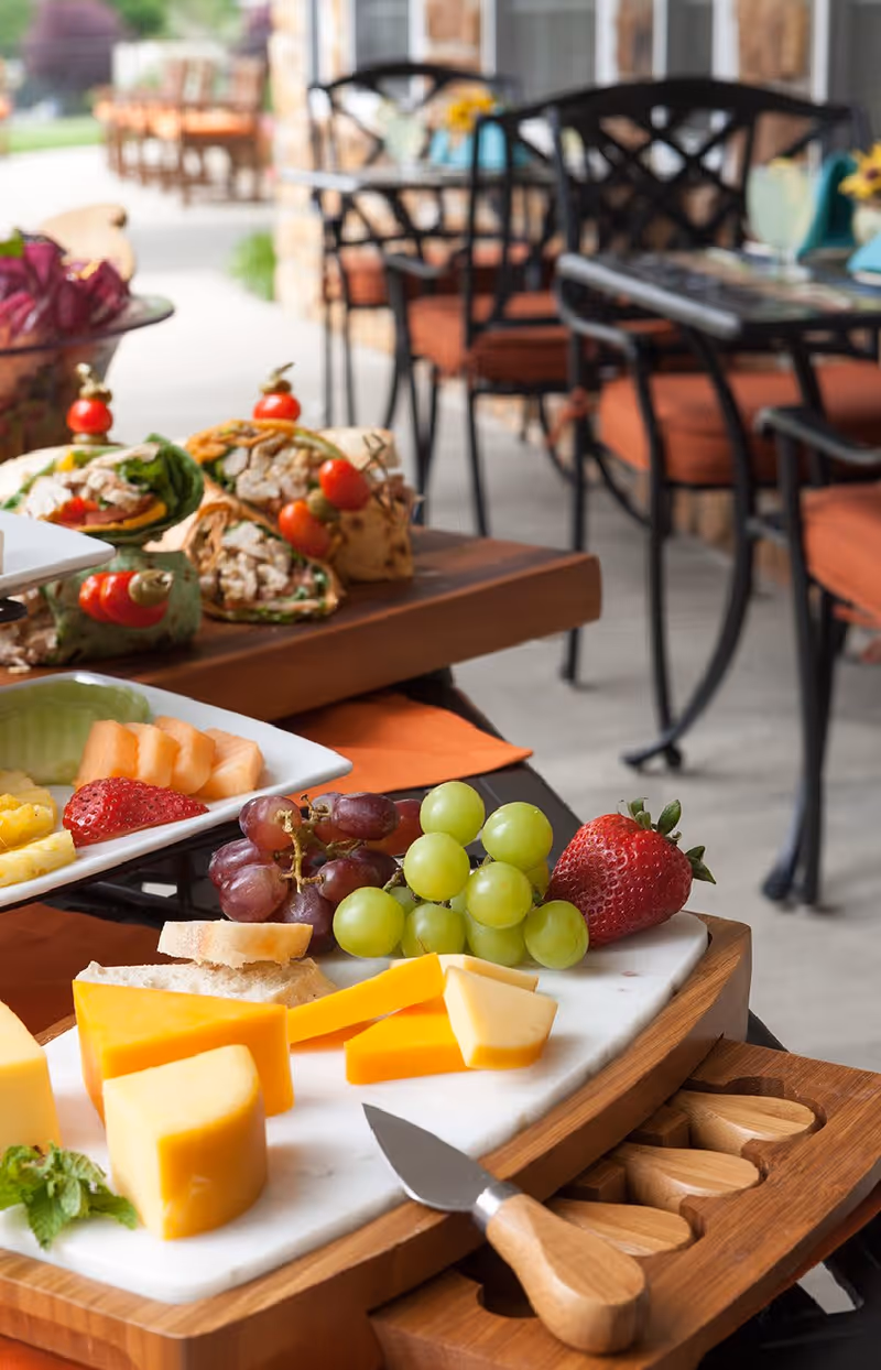 Close-up of a cheese and fruit platter with grapes, strawberries, cantaloupe, and various cheeses on a wooden board with a cheese knife, with wraps and outdoor dining tables and chairs in the background.