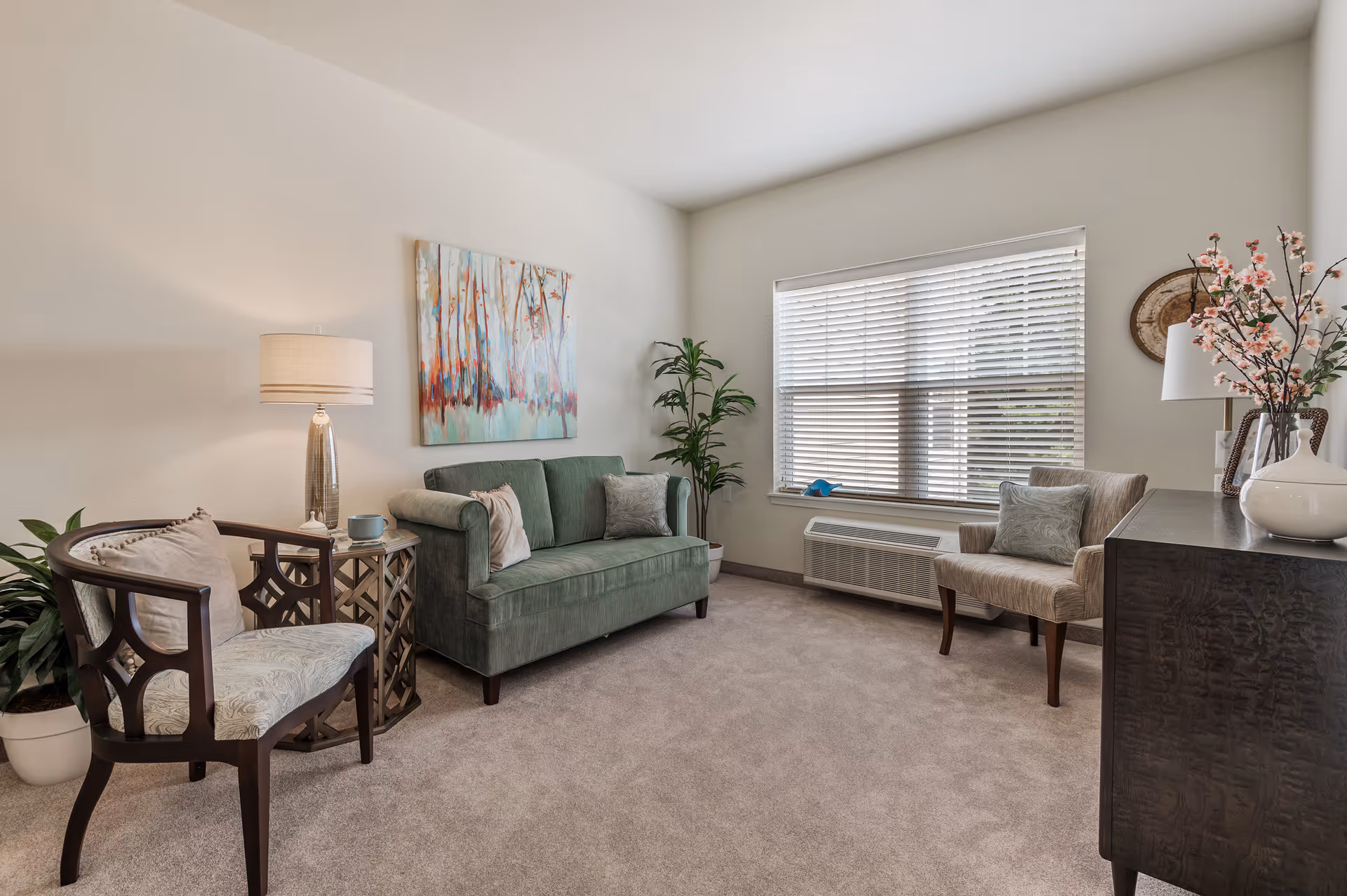 A cozy living room with a green upholstered sofa, two armchairs with patterned cushions, a side table with a lamp and a cup, a large window with blinds, a potted plant, a dark wooden cabinet with decorative items including a vase with flowers, and a colorful abstract painting on the wall.