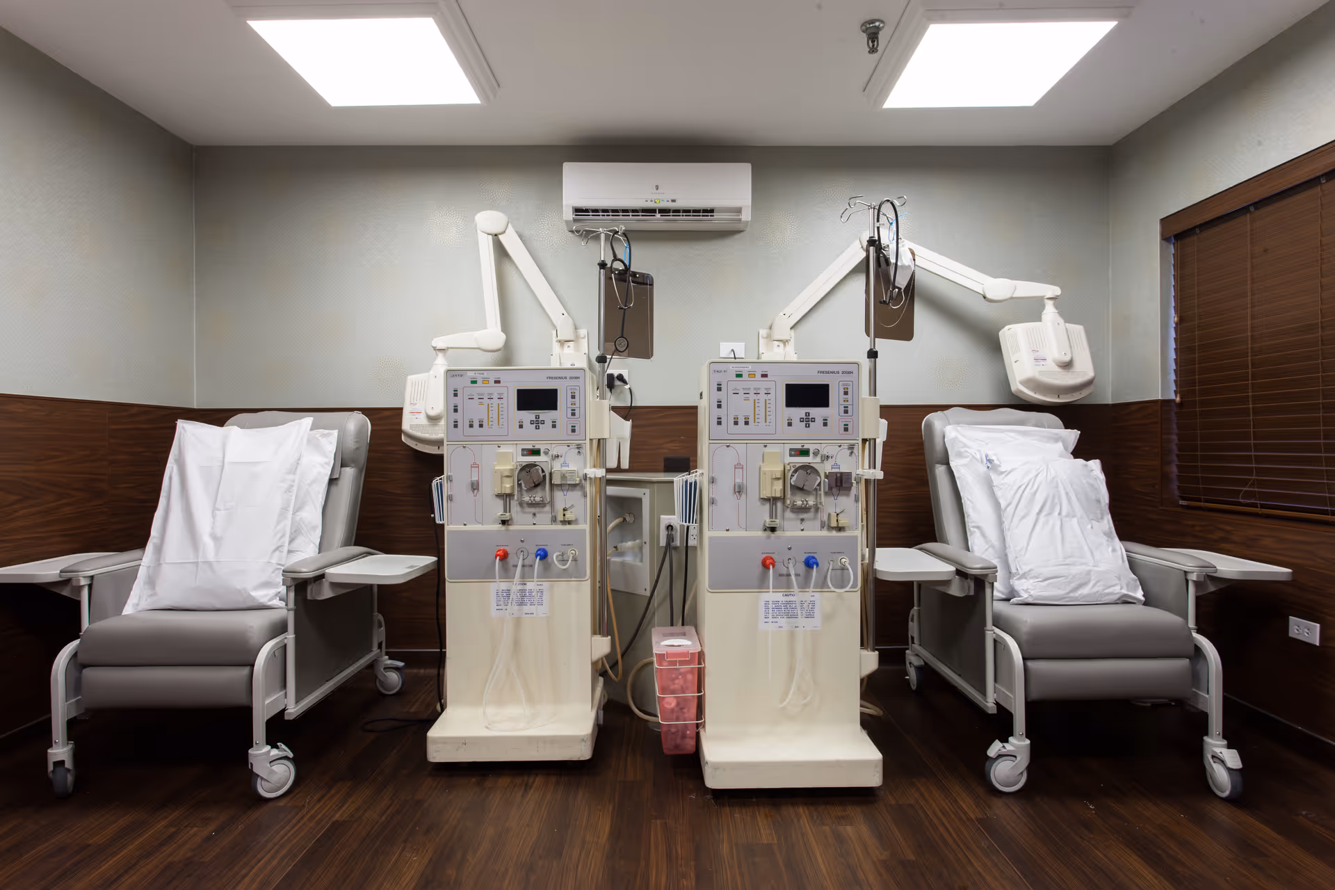 A clinical room with two dialysis machines positioned between two reclining medical chairs with white pillows. The room has wood paneling on the lower half of the walls, a window with closed blinds on the right, and bright overhead lighting.