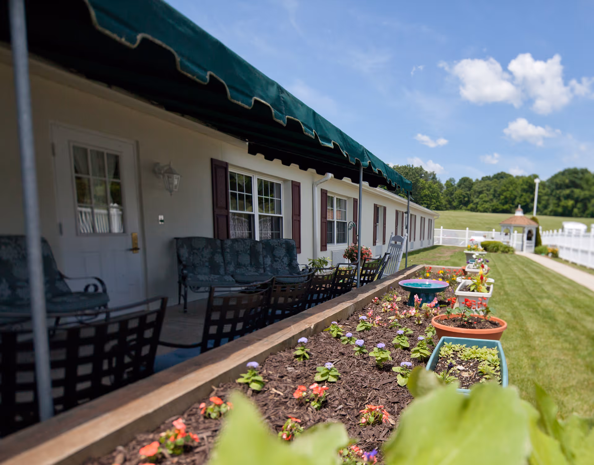 Outdoor patio area of a retirement community with a row of cushioned chairs and tables under a green awning. In the foreground, there is a raised garden bed with various flowers and plants. The background shows a grassy lawn, a white fence, and a small gazebo under a blue sky with scattered clouds.