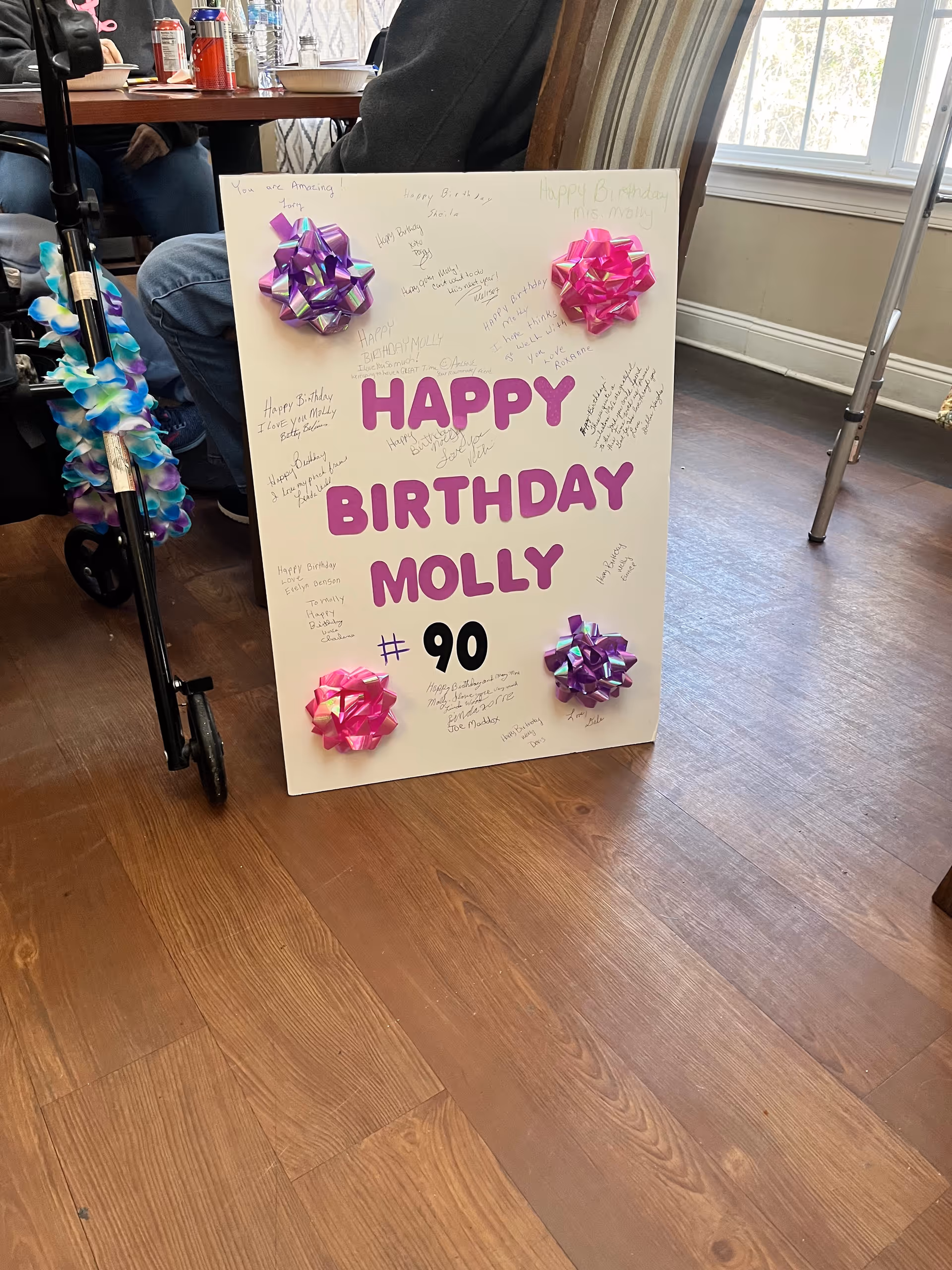 A large decorated poster reading "HAPPY BIRTHDAY MOLLY #90" with colorful bows propped on a wood floor in a communal room next to a walker and a table with drinks.