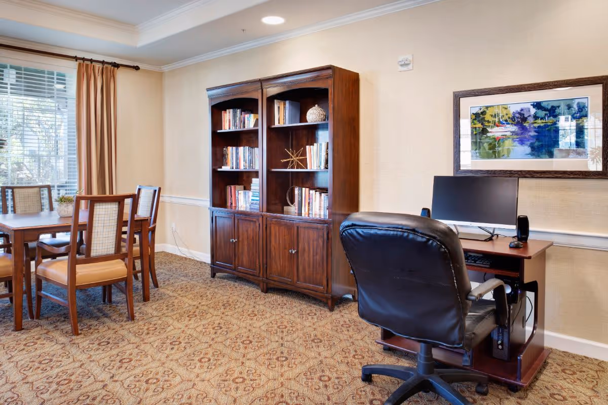 A cozy interior room featuring a wooden bookshelf filled with books, a computer desk with a monitor and black office chair, and a dining table with four chairs near a window with beige curtains. The room has patterned carpet and beige walls with a framed colorful painting above the computer desk.