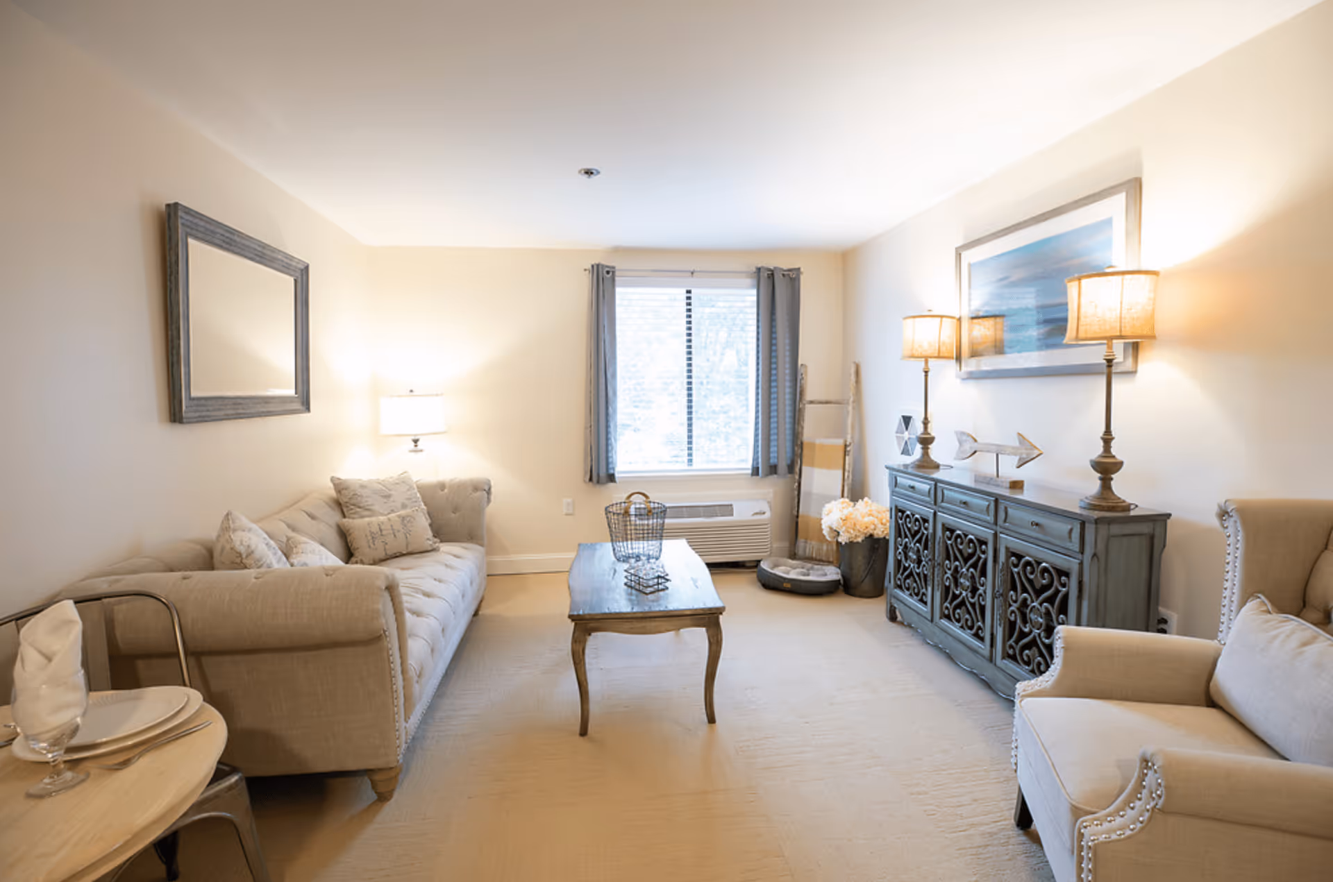 Bright neutral living room with a sofa, armchairs, coffee table, decorative cabinet with lamps, and a window.