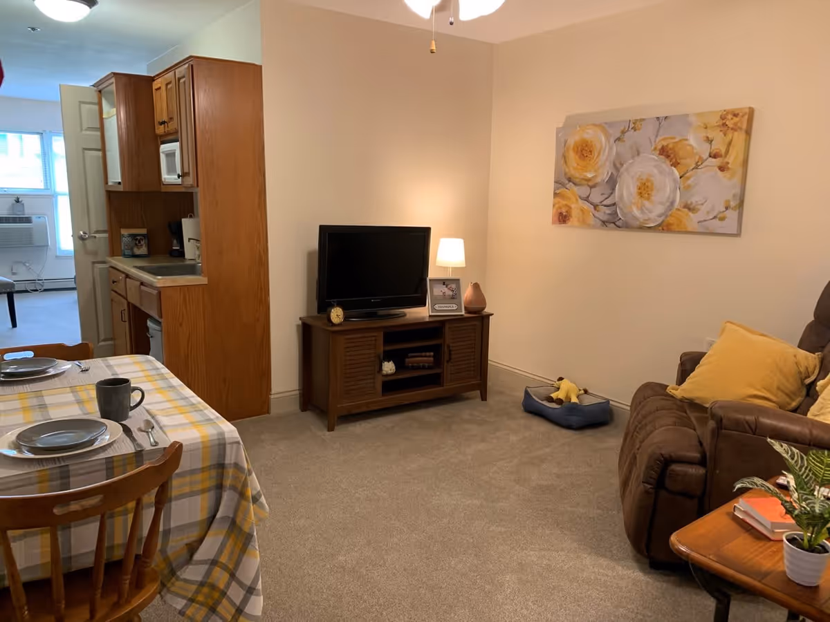 A cozy assisted living apartment living room with a brown recliner sofa adorned with yellow pillows, a wooden TV stand with a flat-screen TV, a small lamp, and decorative items. A dog bed with a toy is on the carpeted floor. To the left, a dining table with a yellow and gray plaid tablecloth is set with plates, utensils, and a mug. The kitchen area with wooden cabinets and a microwave is visible in the background, along with a doorway leading to another room with a window and an air conditioning unit.