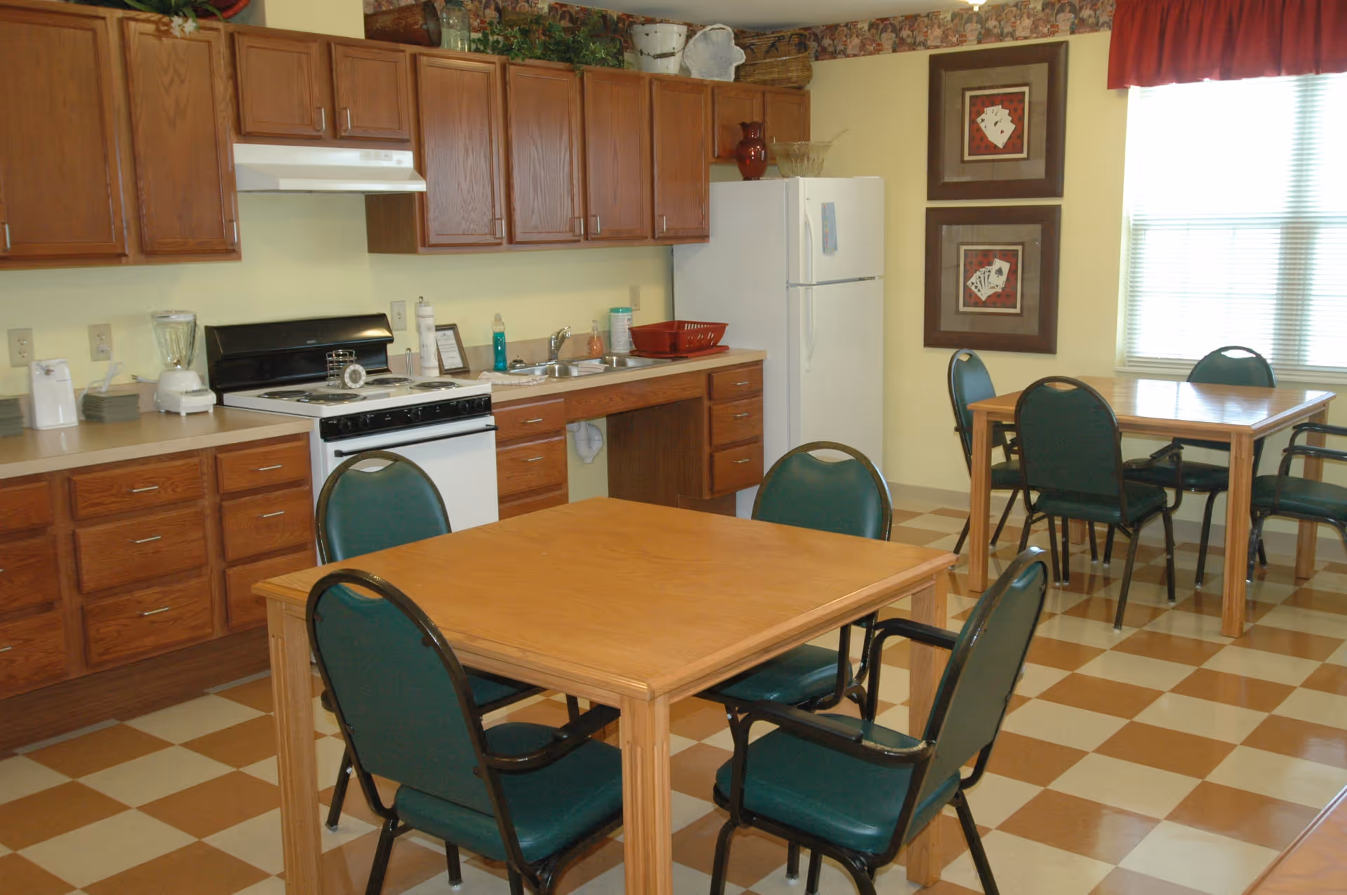Communal dining area with wooden tables and green chairs in front of a kitchenette with wooden cabinets, stove, and refrigerator.