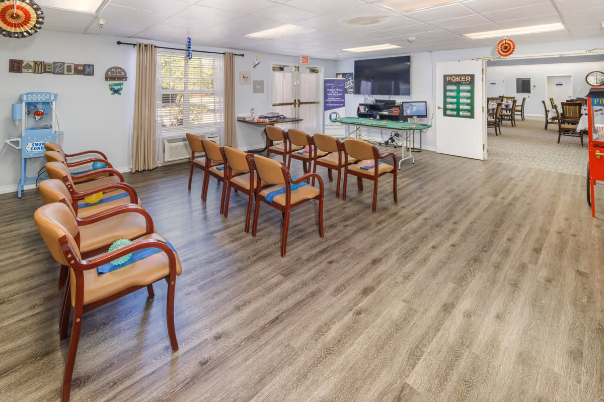 Bright communal activity room with rows of chairs facing a TV and activity tables in an assisted living facility.