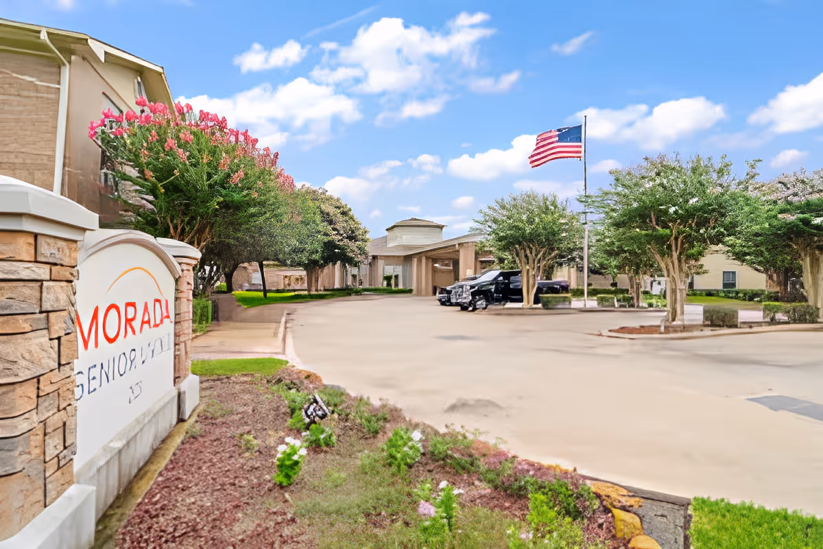 Entrance view of Morada Deer Park senior living facility showing a stone sign with the facility name, a driveway leading to the main building entrance, landscaped flower beds, trees, parked vehicles, and an American flag on a flagpole under a partly cloudy sky.