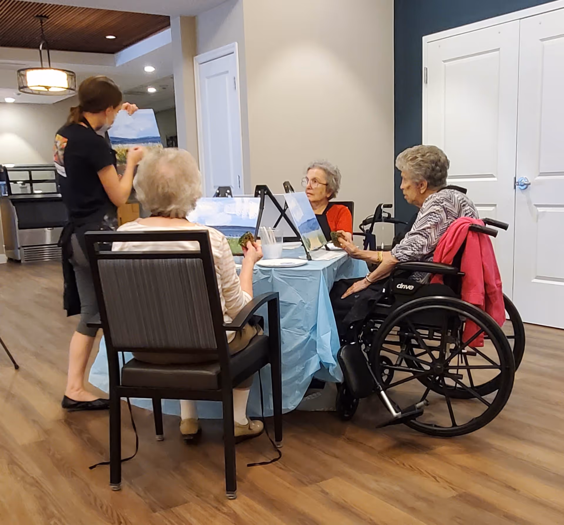 Three elderly women seated around a table covered with a light blue tablecloth, engaged in a painting activity with canvases and paint supplies. One woman is in a wheelchair with a pink jacket draped over the back, another woman is seated in a chair facing away from the camera, and a third woman wearing glasses is looking at her canvas. A younger woman stands nearby holding a painting, possibly instructing or assisting. The room has wooden flooring, white walls, and a ceiling light fixture.